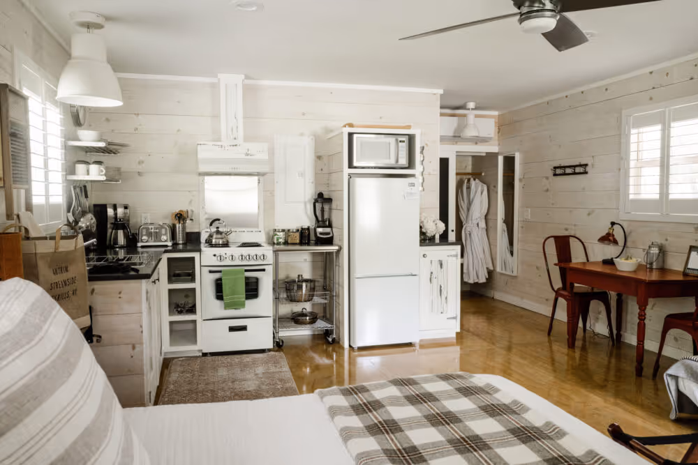 Bright rustic kitchen and dining area with white wooden walls, a white stove, refrigerator, and a small wooden dining table with chairs.