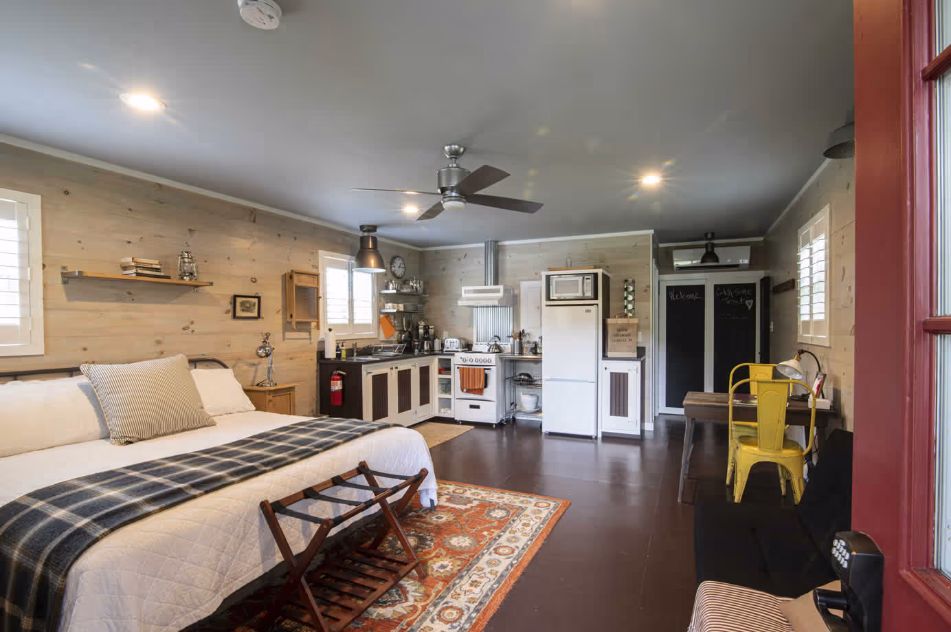 Cozy studio apartment with a bed, plaid blanket, kitchenette, yellow metal chair, desk, ceiling fan, and wooden floor.