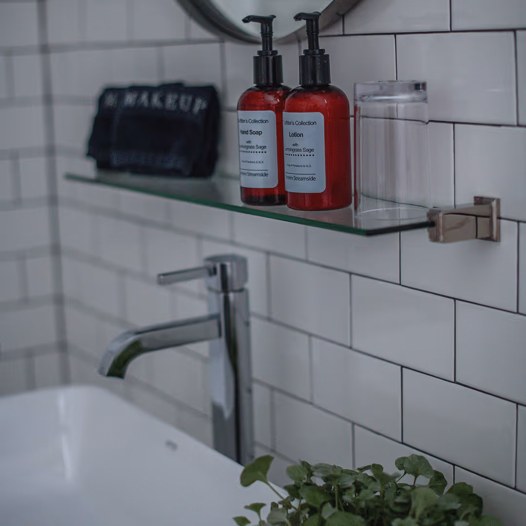 Bathroom sink with chrome faucet, glass shelf holding red soap and lotion dispensers, clear cup, and a small green plant below.