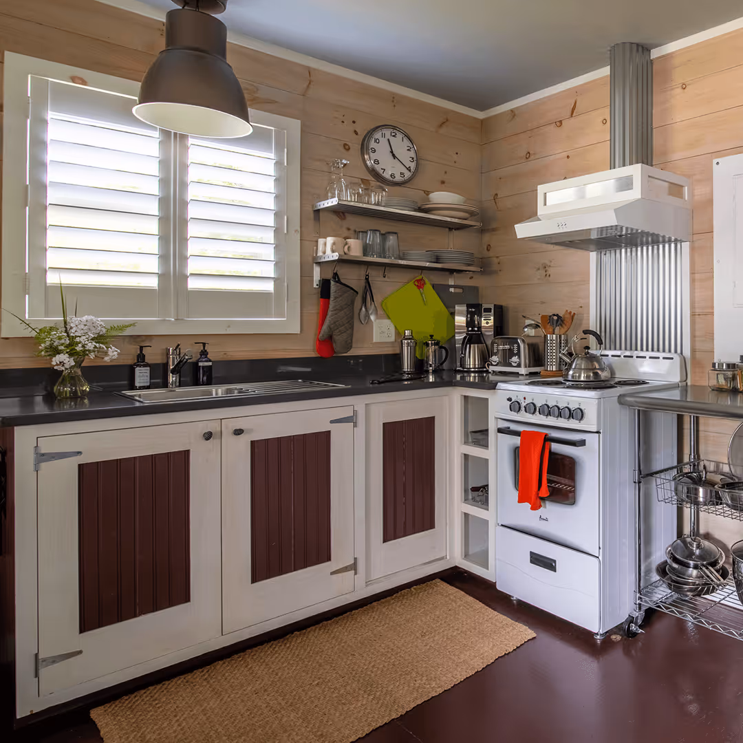 Cozy kitchen with wooden walls, white cabinets with brown panels, black countertop, white stove with red towel, and a window with white shutters.