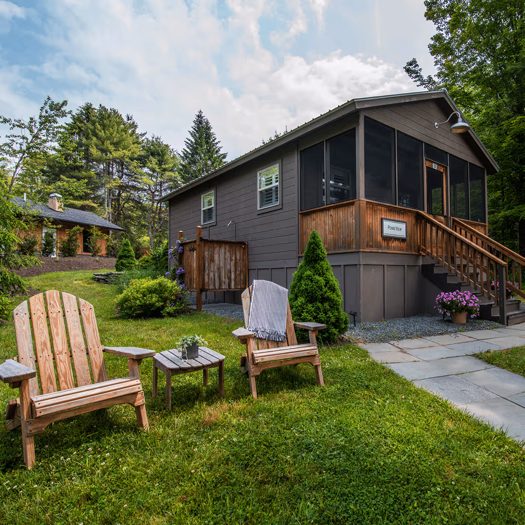 Wooden outdoor chairs and small table on green lawn beside a cabin with screened porch and garden plants.