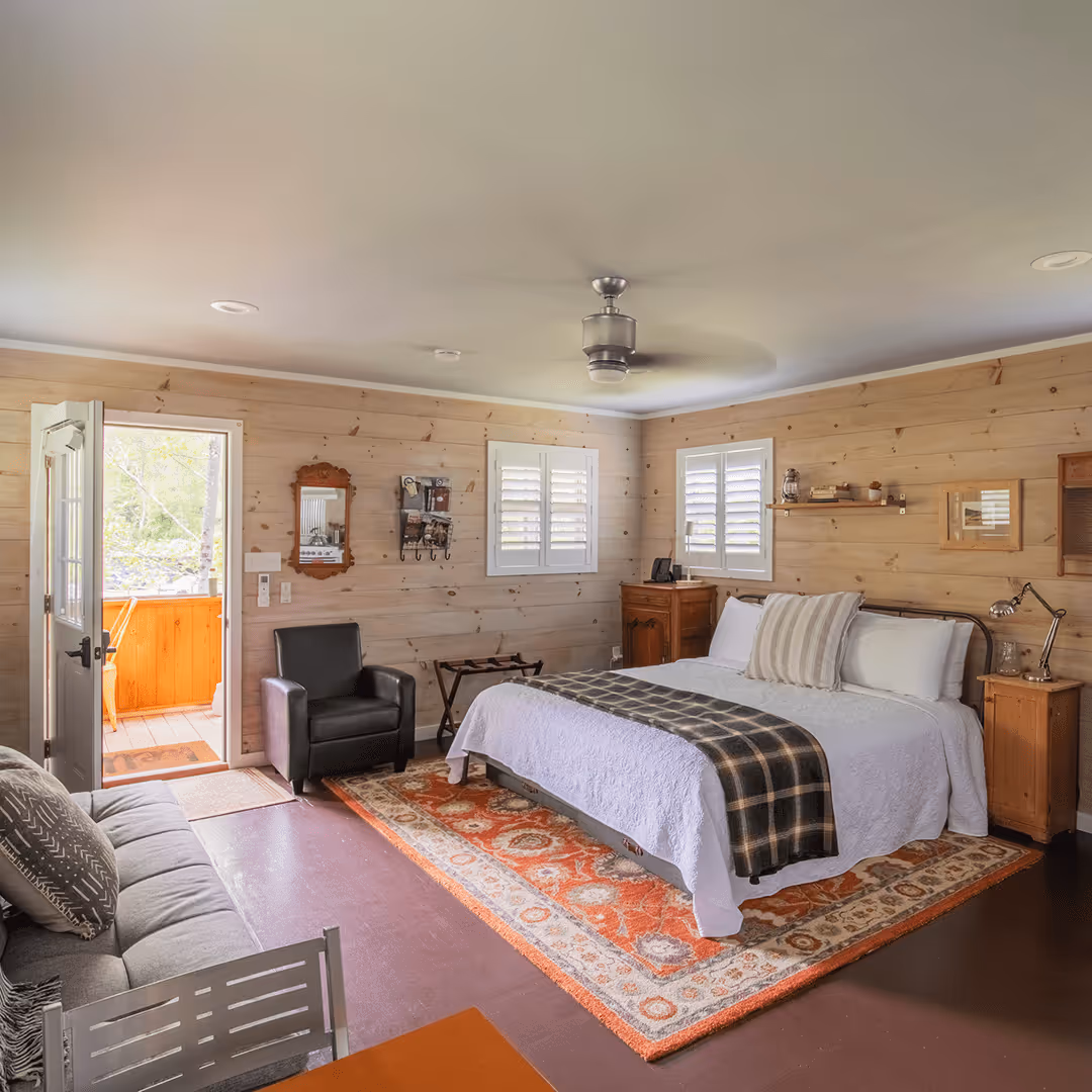 Cozy bedroom with a large bed covered in white and plaid bedding, a black armchair, wooden walls, and an open door leading outside.