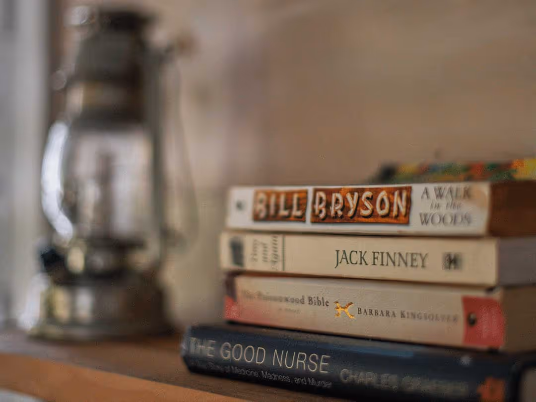 Stack of books on a shelf including titles by Bill Bryson, Jack Finney, Barbara Kingsolver, and Charles on a blurred background with a lantern.