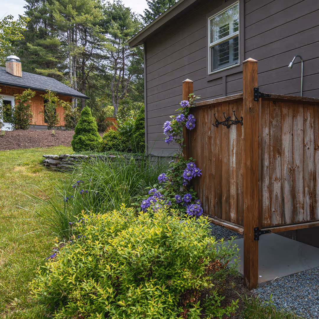 Outdoor wooden shower with hooks and purple flowering vine climbing up the enclosure beside a gray house and green garden.