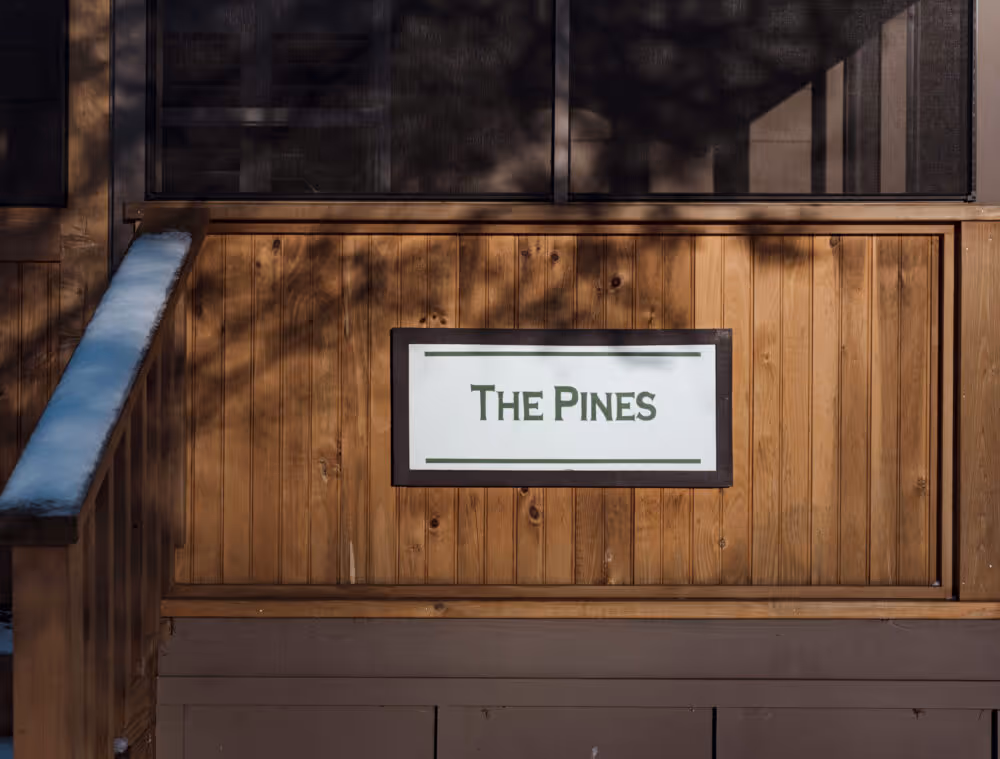 Wooden cabin exterior with a sign reading 'The Pines' mounted on a wooden wall panel beside a snow-dusted railing.