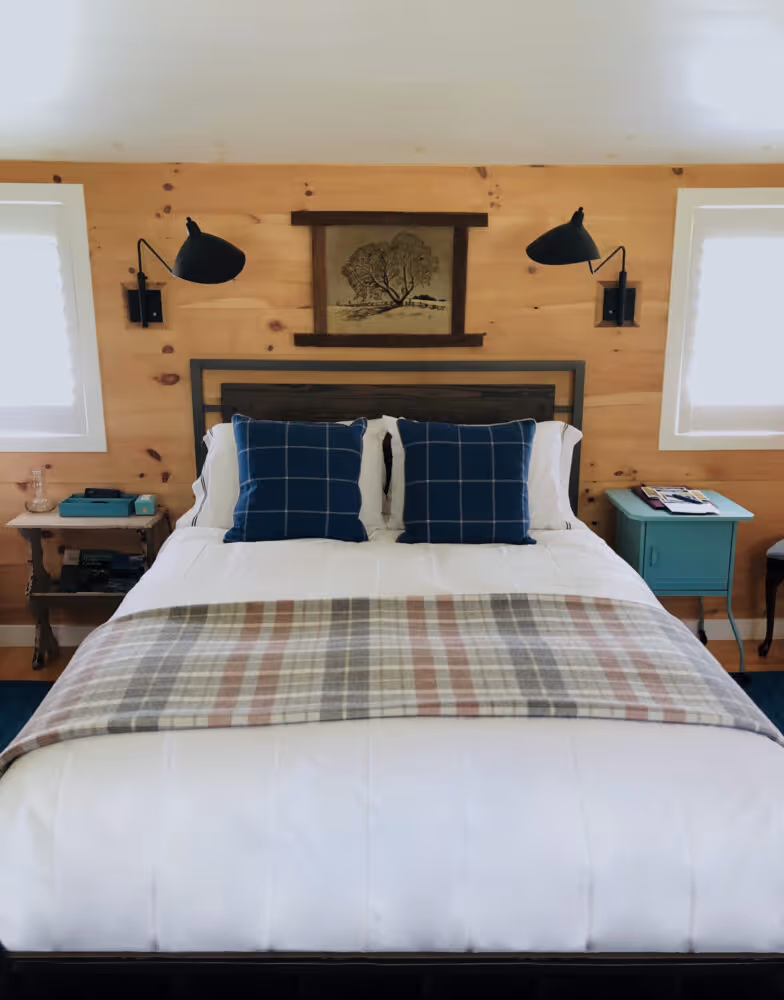 Cozy bedroom with white bedding, plaid throw, two blue checkered pillows, wooden walls, two black wall lamps, and a framed tree artwork.