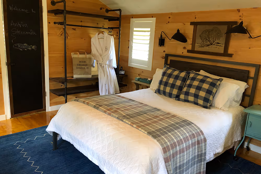Cozy bedroom with wood-paneled walls, a bed with white bedding and plaid pillows, a hanging white robe, and a chalkboard with 'Welcome to Antrim Streamside' written on it.