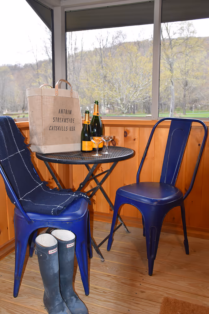 Two blue metal chairs with a blanket on one, a small round black table with bottles of sparkling wine and two coupe glasses, beige tote bag with 'ANTRIM STREAMSIDE CATSKILLS USA' text, and black Hunter rain boots on wooden floor in a screened porch.