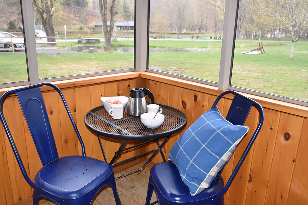 Small round metal table with coffee pot, two cups, a bowl of apples, and a bowl with a spoon, between two blue metal chairs, one with a blue plaid pillow, in a wooden screened porch overlooking a green yard.