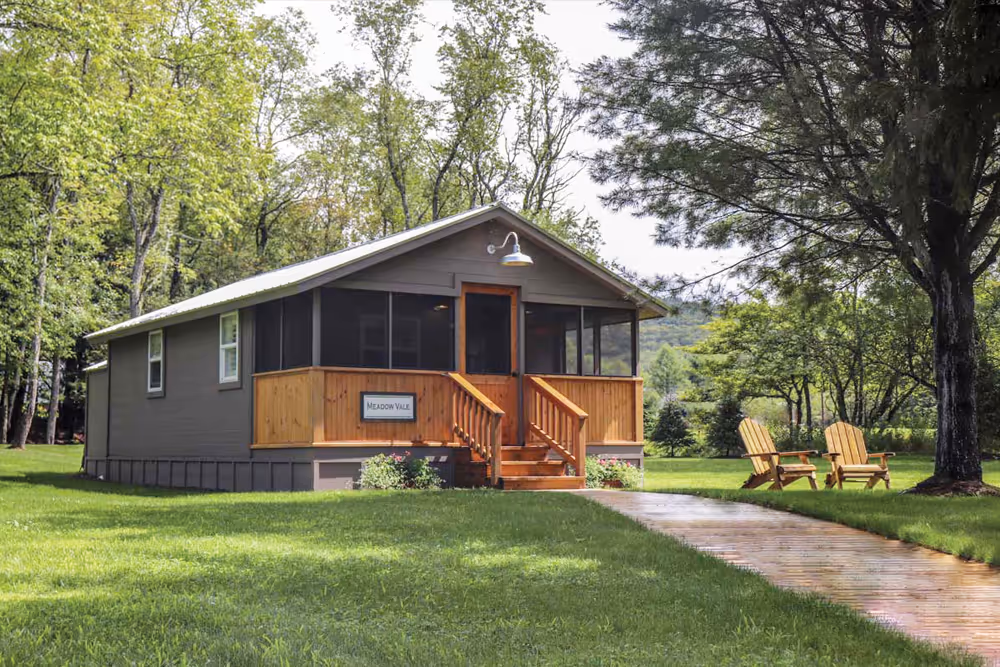Small gray cabin with wooden porch and steps on green lawn surrounded by trees, with two wooden chairs near a large tree.