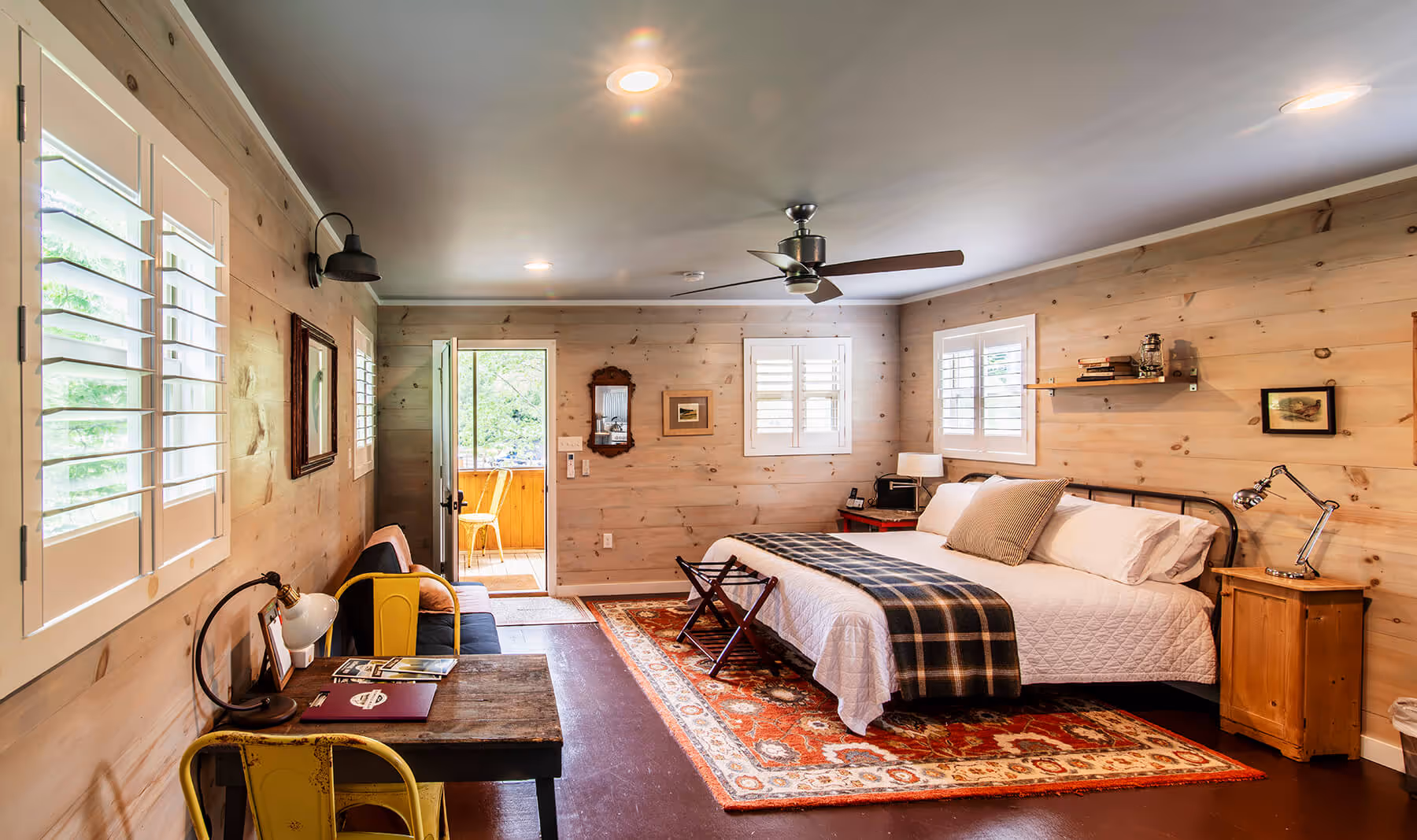 Rustic bedroom with wood-paneled walls, a large bed with white and plaid bedding, a red patterned rug, desk with yellow chairs, and a door leading to a porch.