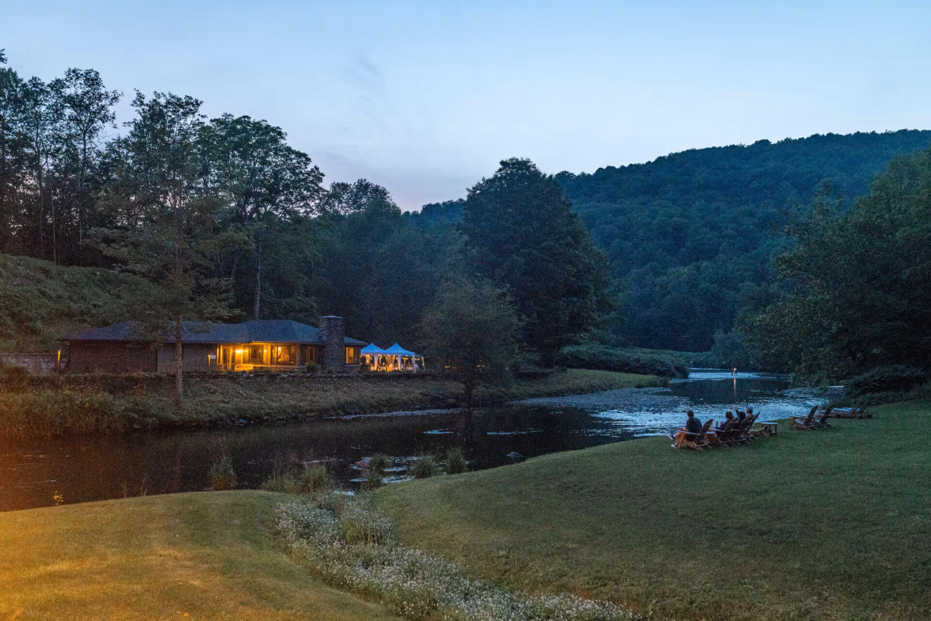 Evening view of a riverside house with glowing lights, surrounded by trees and hills, with people sitting on chairs near the water.