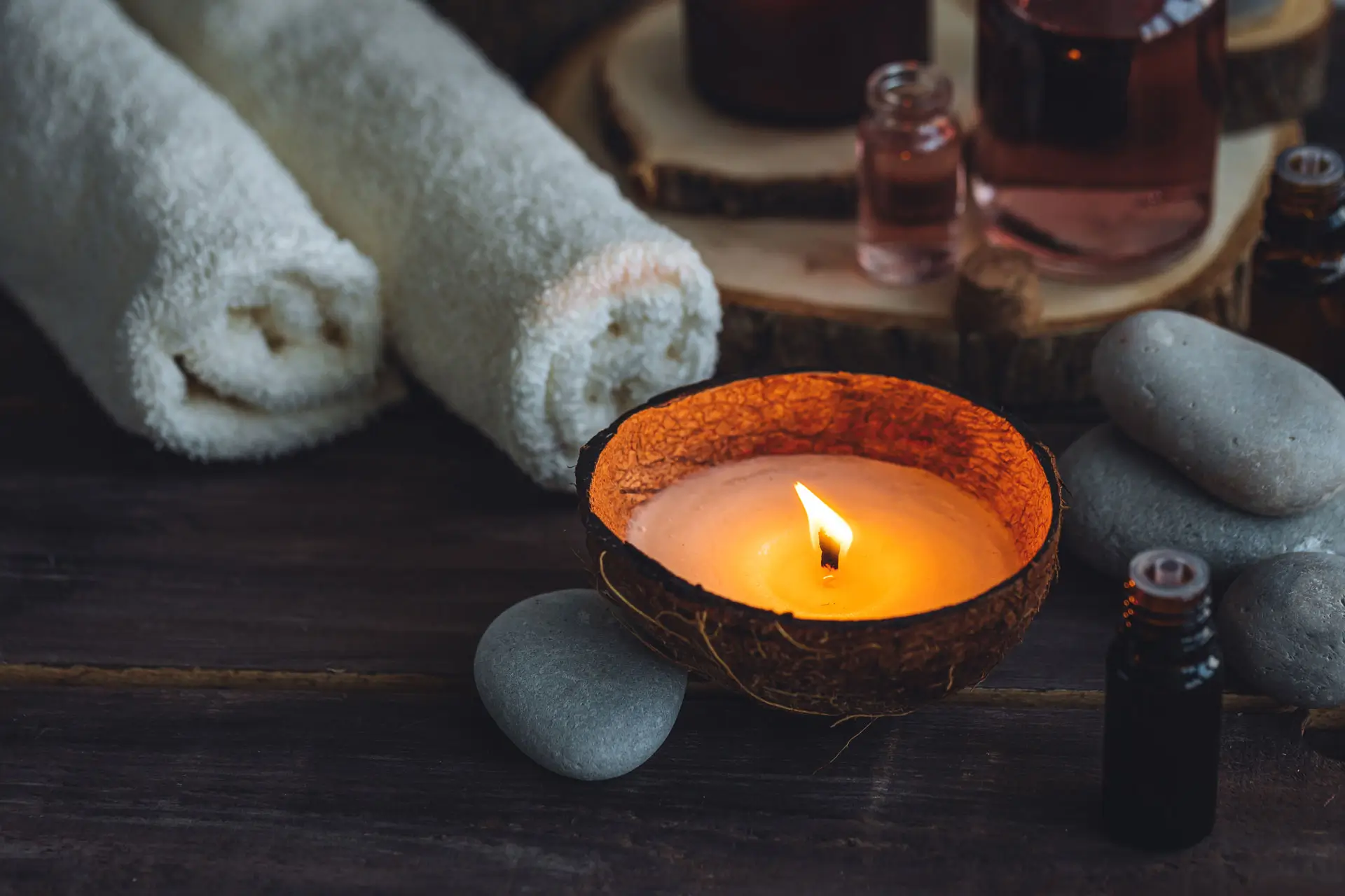 Lit candle in a coconut shell surrounded by smooth stones, rolled white towels, and small bottles on a dark wooden surface.