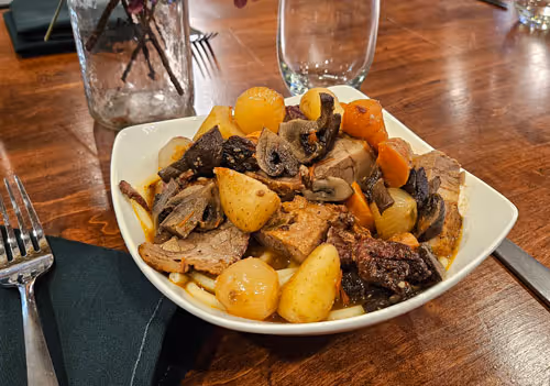 Bowl of pot roast with sliced beef, mushrooms, baby potatoes, carrots, and pearl onions on a wooden table.