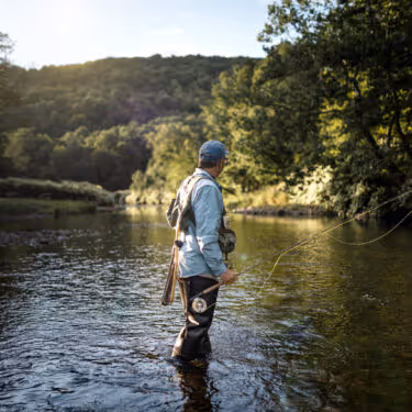 Man fly fishing in a sunlit river surrounded by green forest hills.