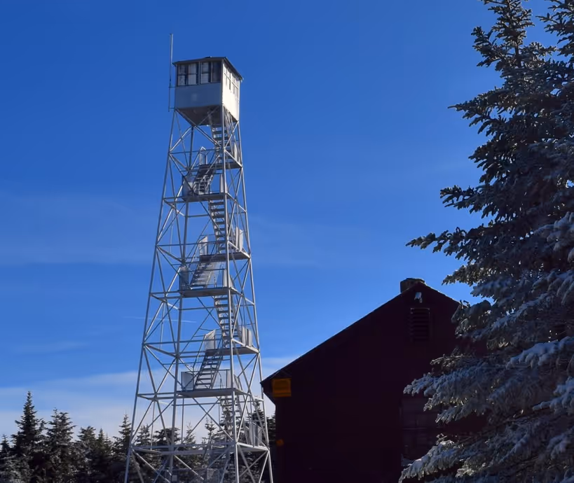 Metal fire lookout tower with stairs and an observation cabin at the top next to a building and pine trees under a clear blue sky.