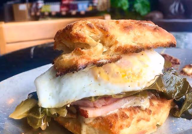 Breakfast sandwich with a fried egg, ham, and leafy greens on toasted bread on a plate.