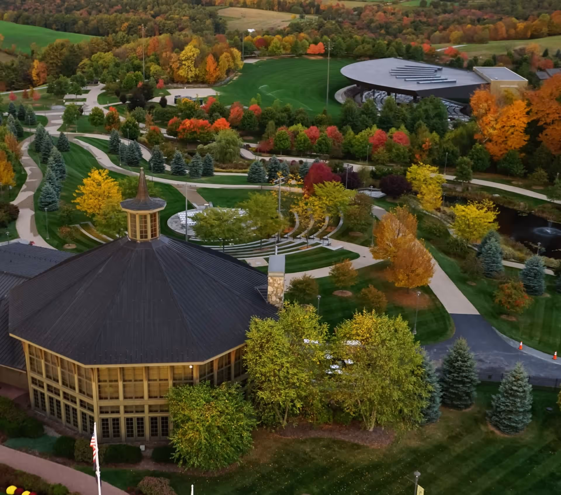 Aerial view of a park in autumn with colorful trees, winding paved paths, large octagonal building with a dark roof, and a modern amphitheater.