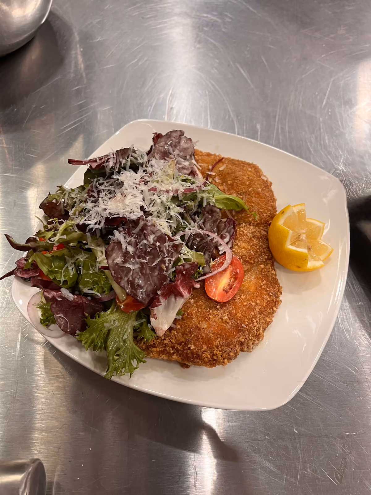 Breaded and fried cutlets served with mixed green salad topped with shredded cheese and a lemon wedge on a white plate.