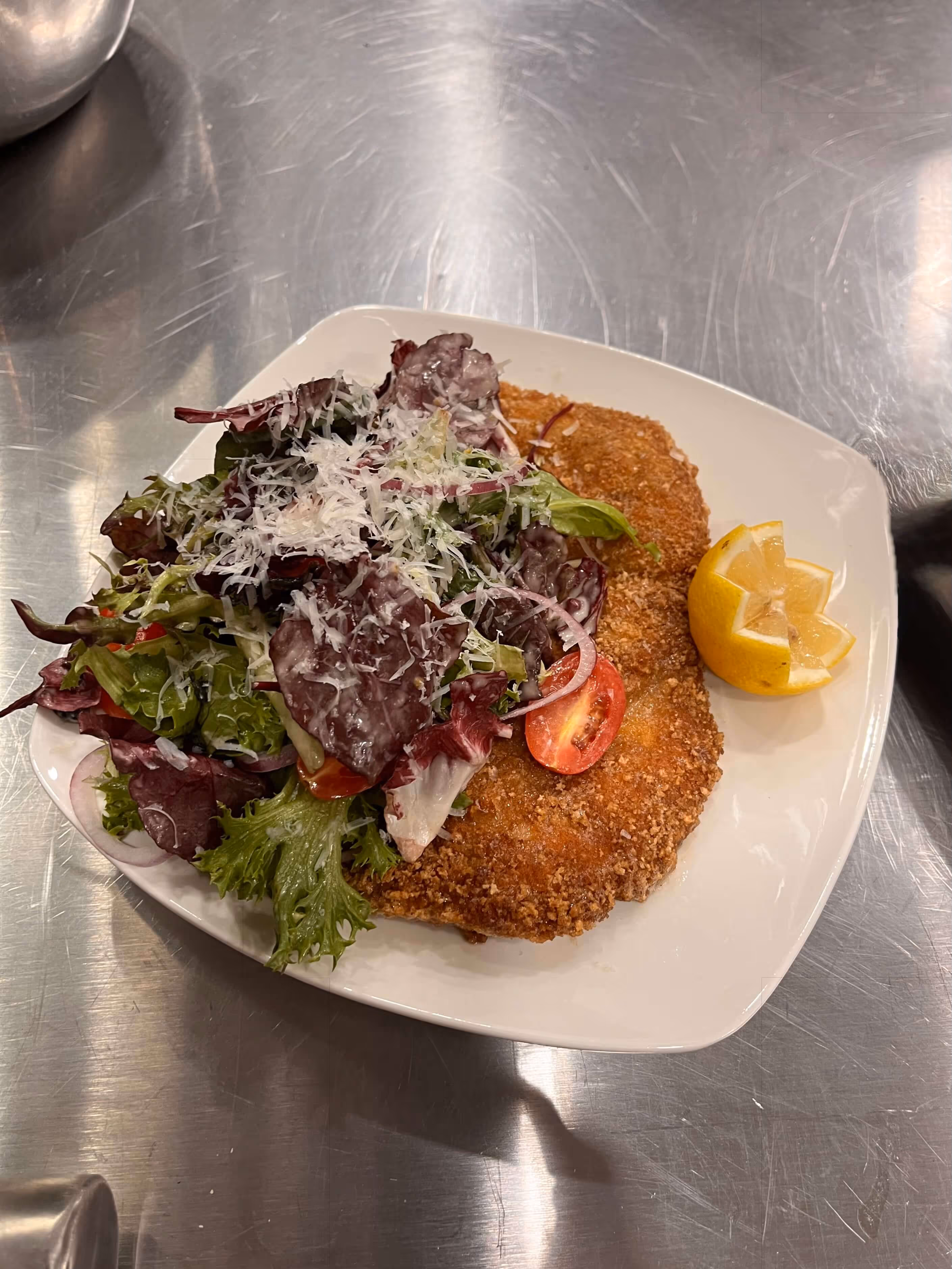 Breaded and fried cutlets served with mixed green salad topped with shredded cheese and a lemon wedge on a white plate.