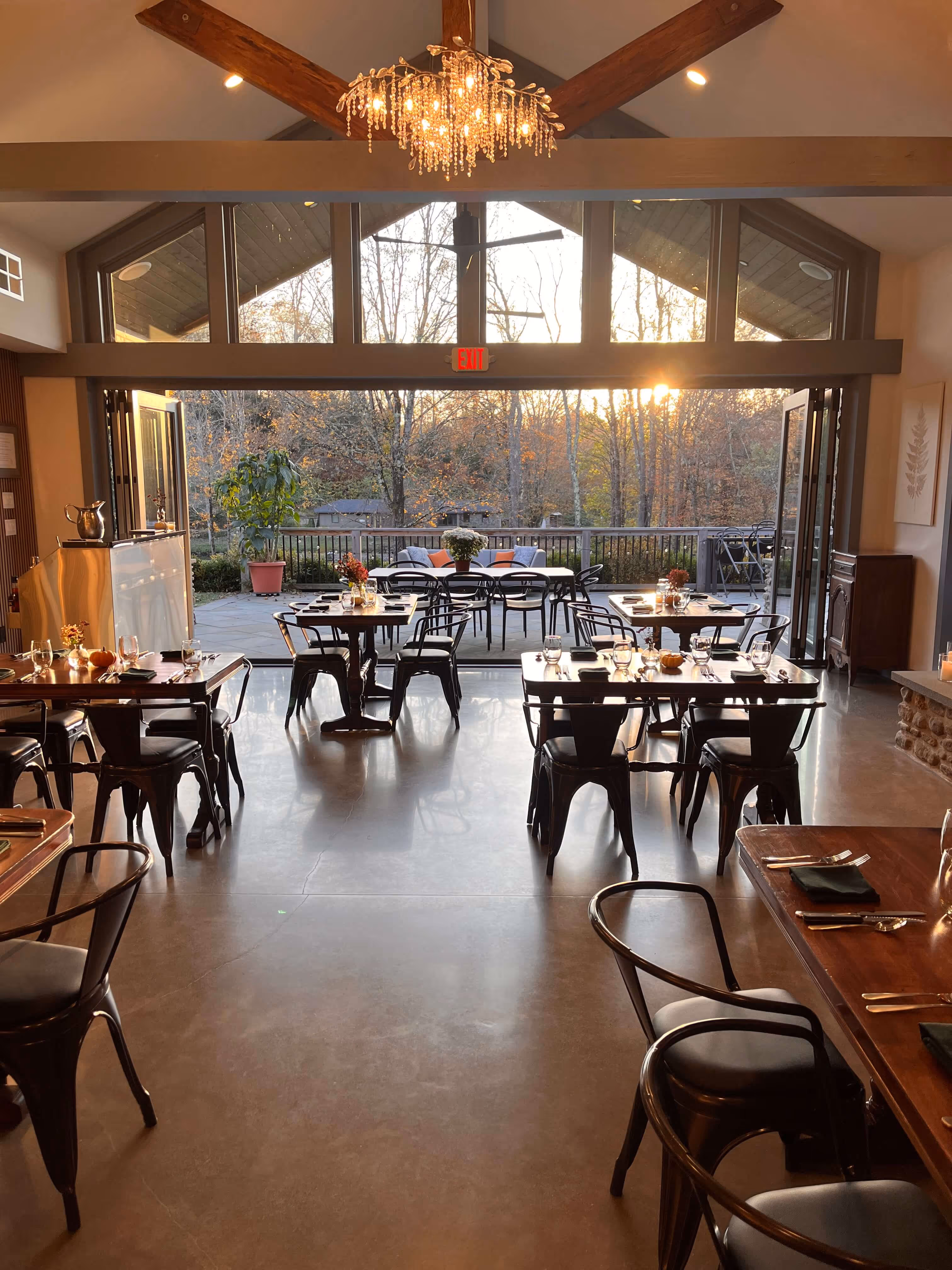 Cozy restaurant dining area with wooden tables and black metal chairs set for guests, leading to an outdoor seating area with trees and sunset in the background.