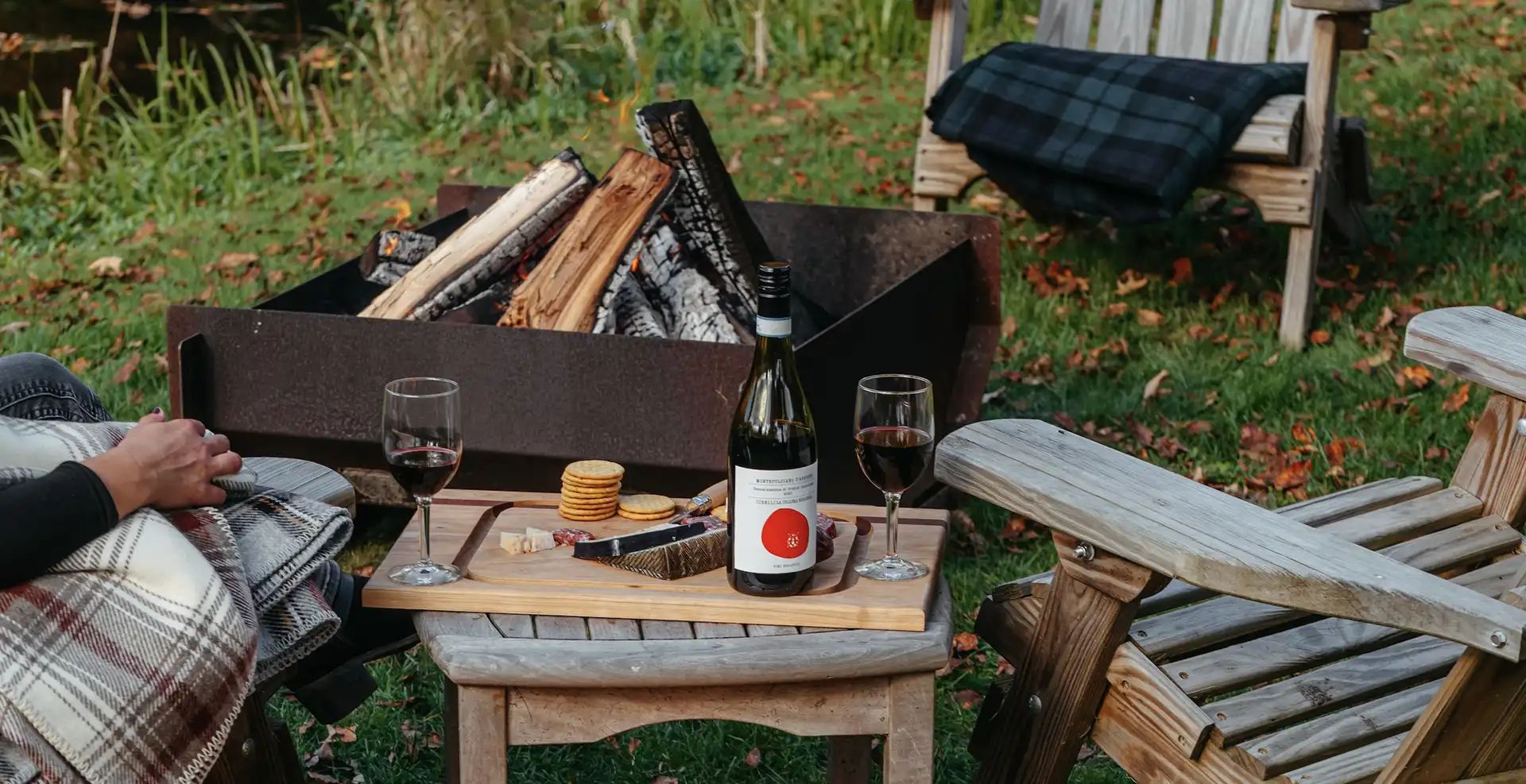 Outdoor scene with a fire pit burning wood, two wooden chairs with blankets, a small table holding a wine bottle, two glasses of red wine, and a snack tray with crackers, cheese, and sausage.