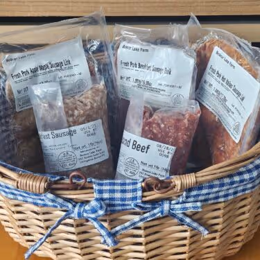 Wicker basket with several packaged meats labeled as fresh pork, beef sausage, and ground beef with a blue and white checkered cloth bow.