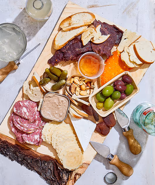 Charcuterie board with sliced salami, cheese wedges, crackers, pickles, olives, nuts, bread slices, and a small container of honey, accompanied by two spreader knives and drinks.