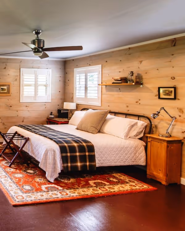 Cozy bedroom with wood-paneled walls, white bed with plaid throw, colorful rug, bedside tables, and ceiling fan.