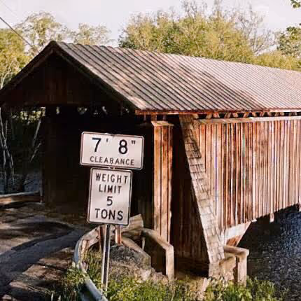 Wooden covered bridge over a small body of water with clearance and weight limit signs at the entrance.