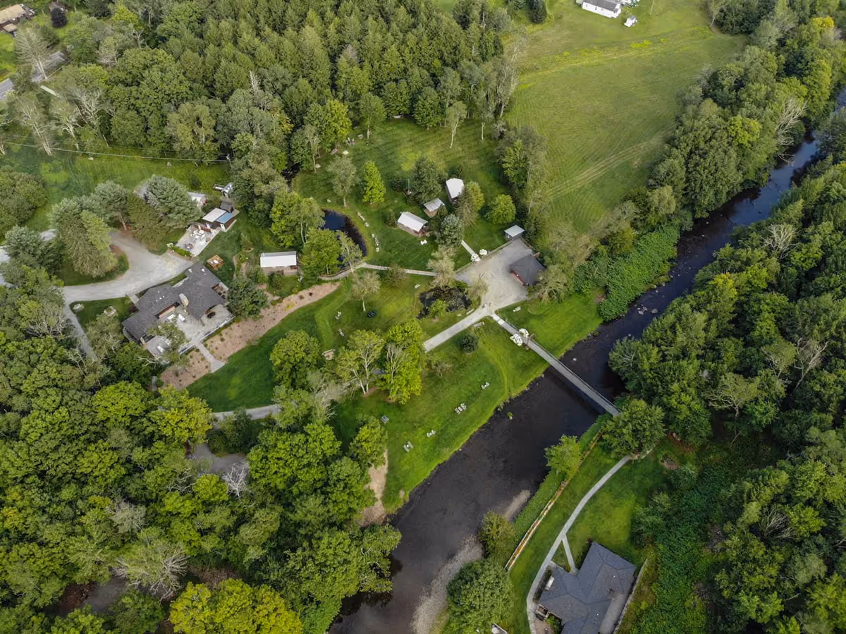 Aerial view of green landscape with house, cabins, pond, wooded areas, and a river with footbridges.
