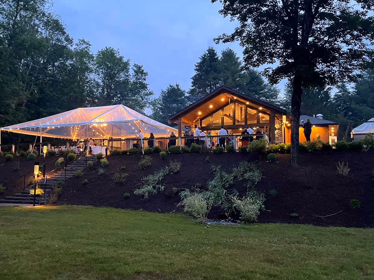 Evening view of a lit wooden lodge with people on the deck near a clear tent in a forest setting.