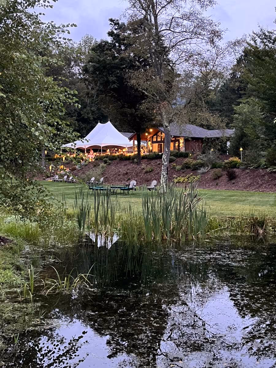 Lit white event tent and chairs on grassy hill reflecting in a pond at dusk surrounded by trees.