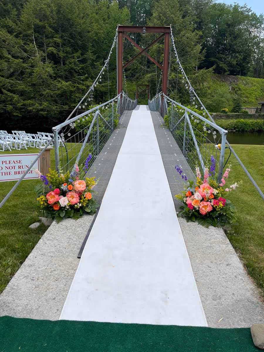 Suspension bridge with white runner and colorful floral arrangements on each side, surrounded by green trees.