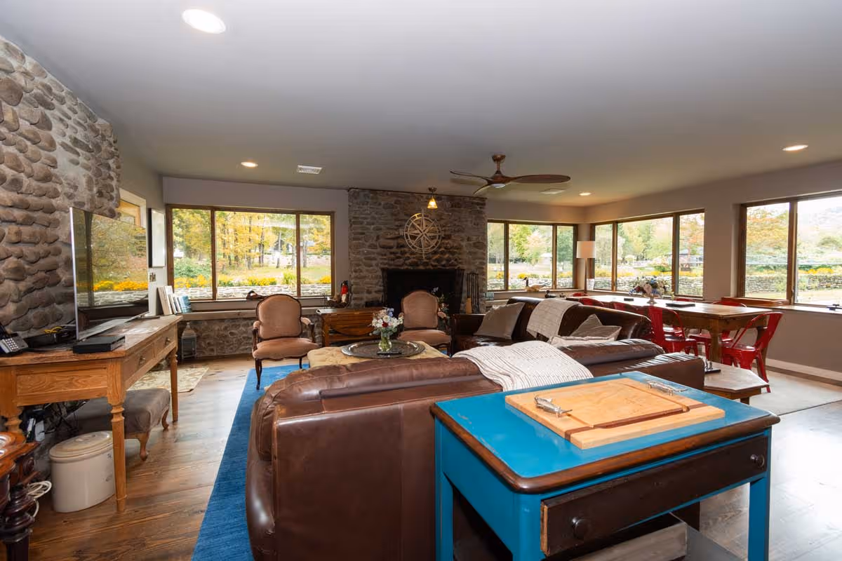 Cozy living room with stone fireplace, large windows, leather sofas, wooden furniture, and blue rug.