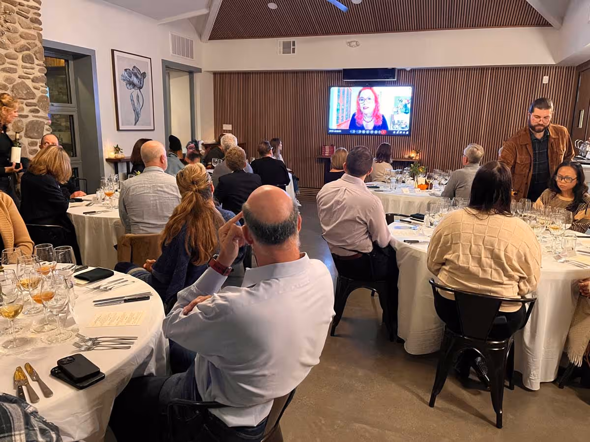People seated at round tables watching a woman speaking on a large screen in a modern room.