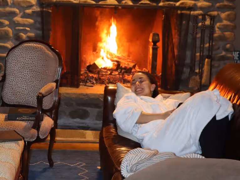 Woman in white robe smiling on brown couch near lit stone fireplace with armchair and book nearby.