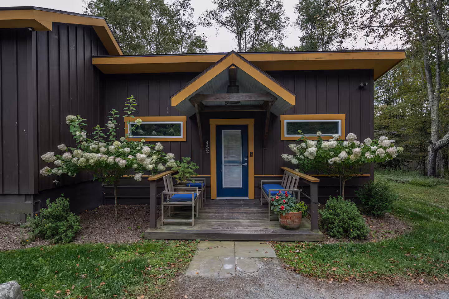 Dark brown house entrance with yellow trim, blue door, porch with chairs, and white flowering bushes.