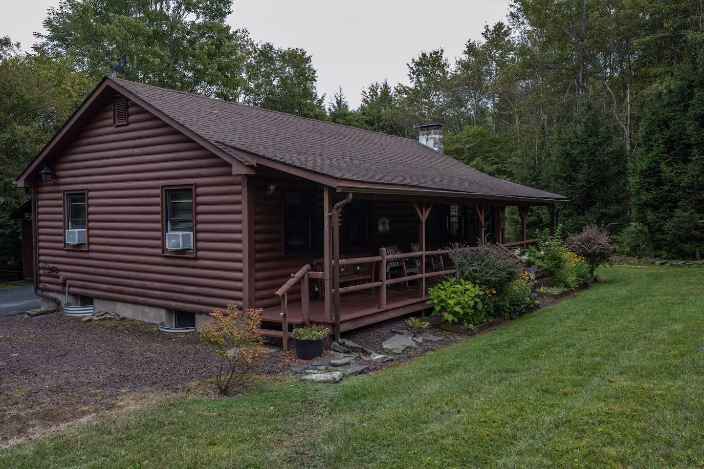 Brown wooden cabin with a porch and railing, surrounded by green plants and lawn, in a forest setting.