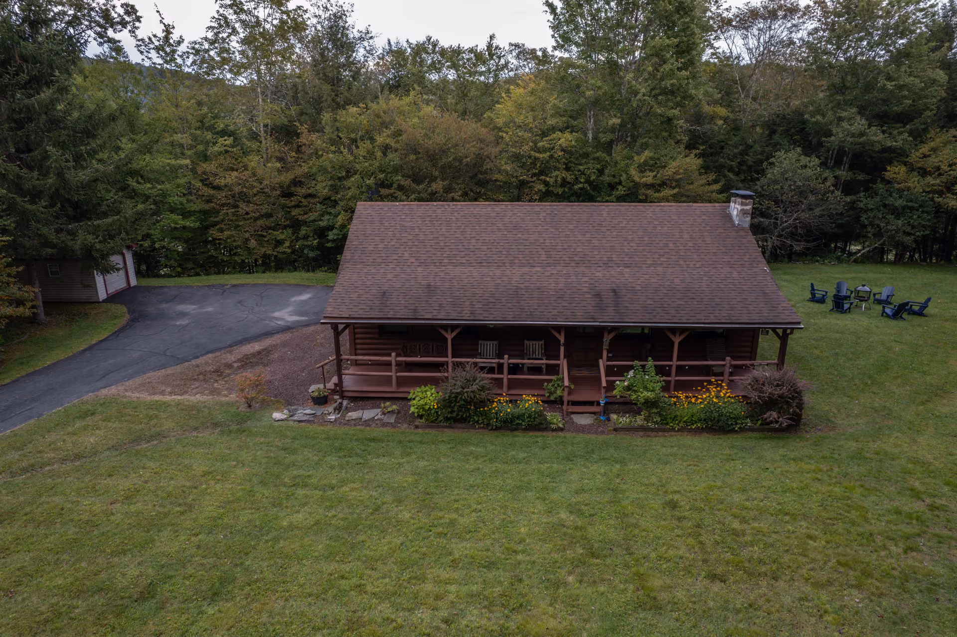 Wooden cabin with a porch and chairs surrounded by green lawn and trees in the background.