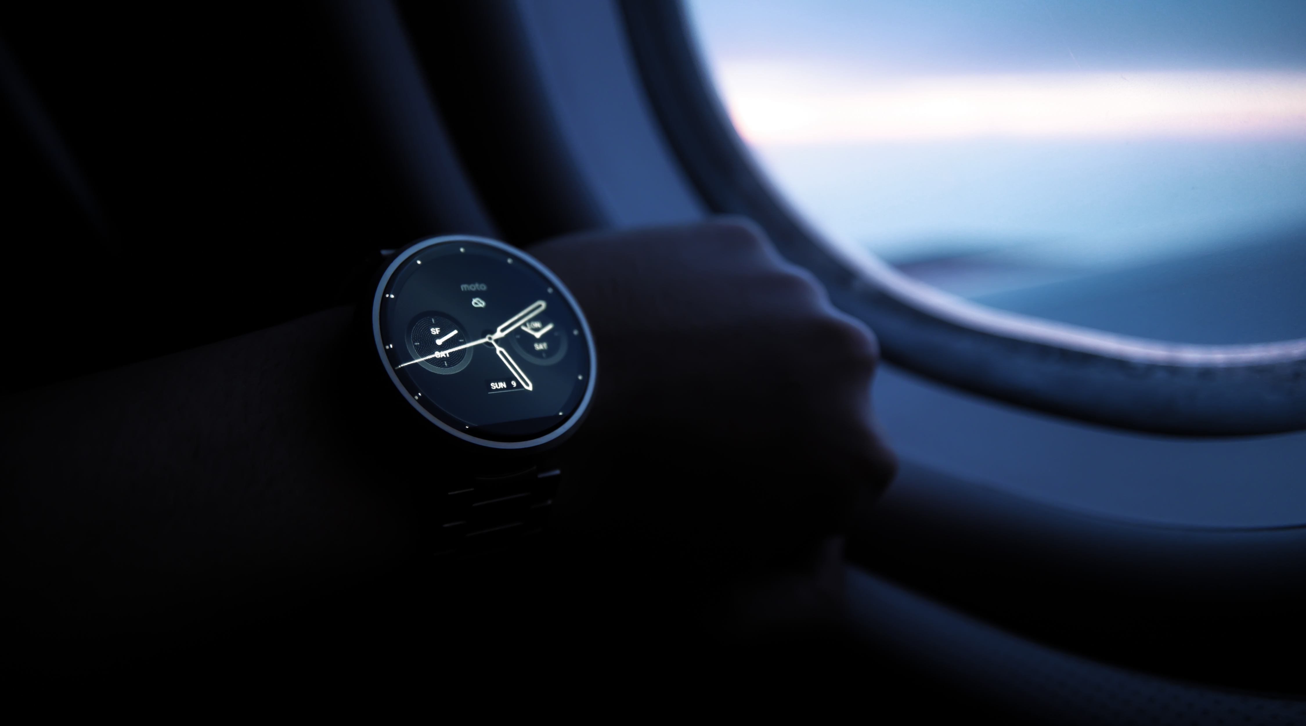 Round-faced smartwatch worn on a wrist beside an aeroplane window