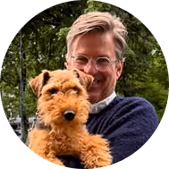 Man wearing glasses holds a small brown dog outdoors with trees in the background.