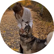 Woman in white coat smiling while petting a large dog on a leaf-covered path.