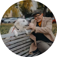 Smiling man wearing sunglasses holding a small fluffy dog outdoors with blue sky background.