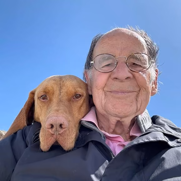 Elderly man with glasses smiling closely with brown dog under bright blue sky.