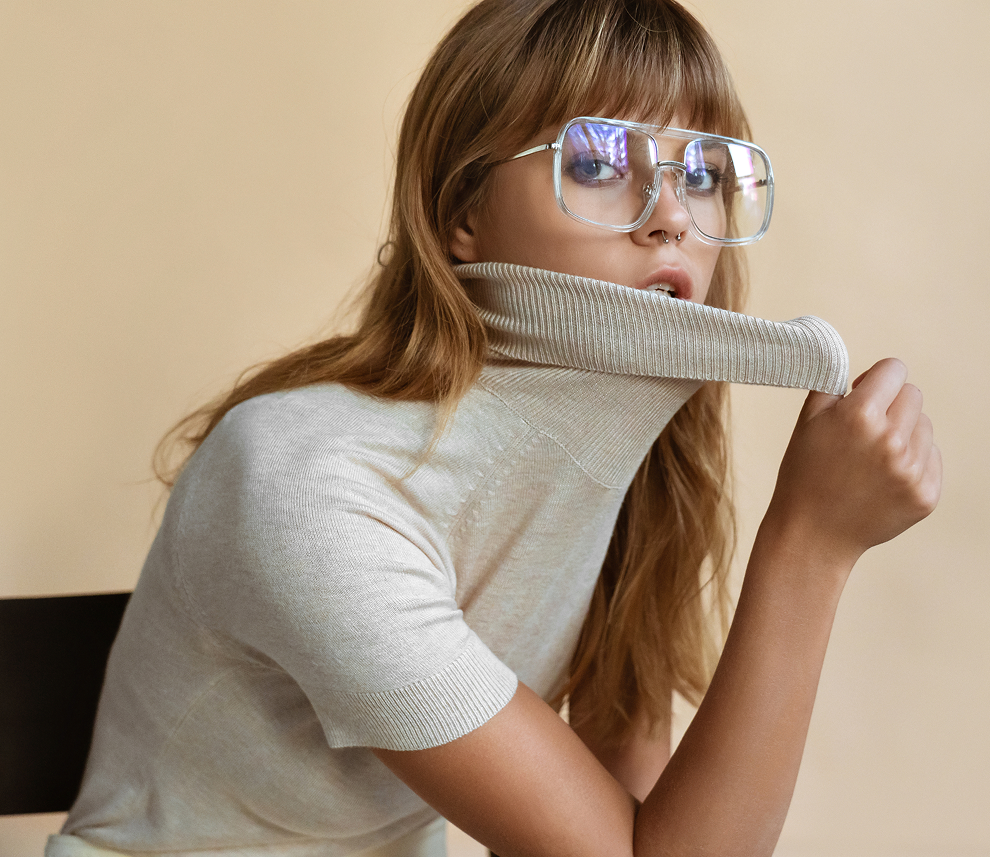 Young woman with light brown hair wearing large clear glasses and a beige short-sleeve turtleneck sweater, pulling the collar over her mouth.