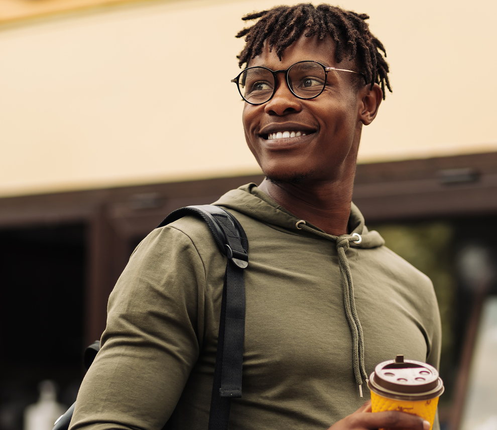 Smiling man with glasses wearing a green hoodie and carrying a backpack, holding a yellow coffee cup.