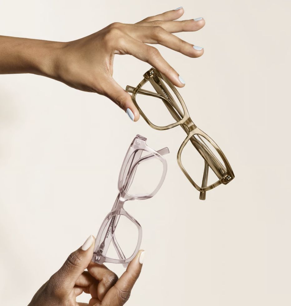 Two hands holding eyeglasses with translucent frames, one in brown and the other in light pink, against a neutral background.