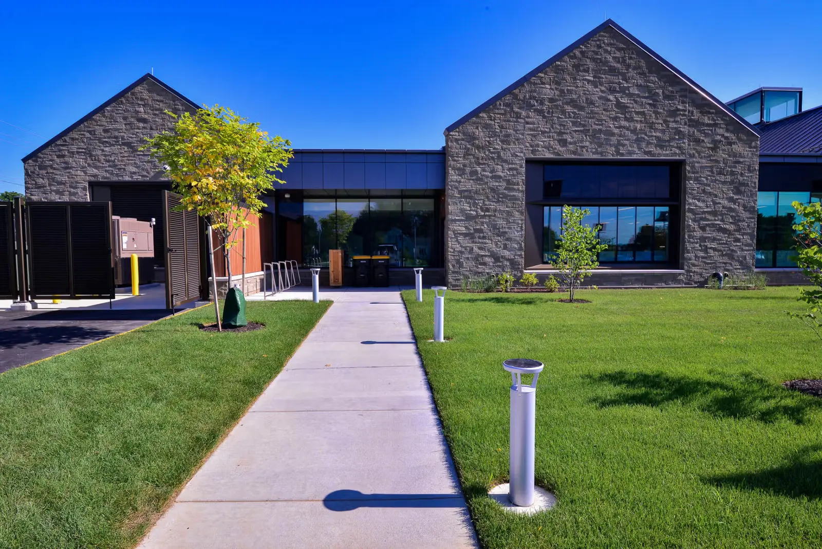 Modern commercial building with stone facades, large windows, and a concrete walkway surrounded by green grass and small trees.