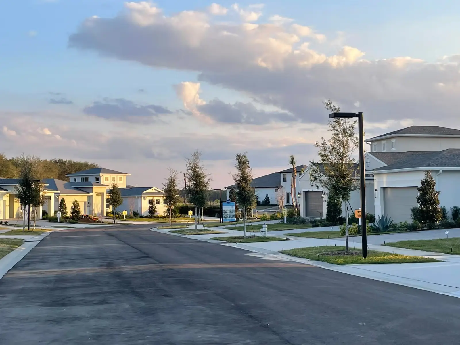 Quiet residential street with modern houses, young trees, and street lamps under a partly cloudy sky at sunset.